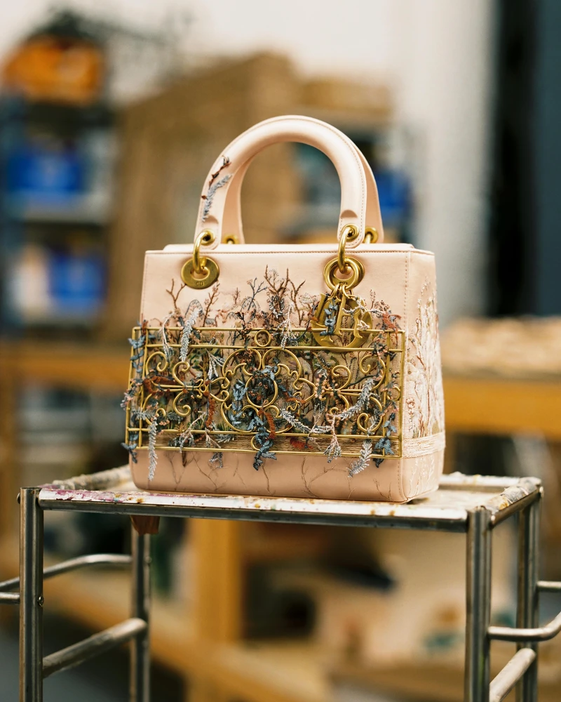 Dior Lady Art Eva Jospin 中型提包<，以米色緞面製成，於吊飾周圍以交錯絲線刺繡點綴，並飾有一座象徵蒙田大道30號總店陽台的金屬陽台造型。搭配古金色金屬飾件。© Marion Berrin。